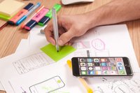 Hand, die mit einem Stift auf einen grünen Zettel schreibt. Daneben liegen Smartphone und Blätter mit Skizzen.